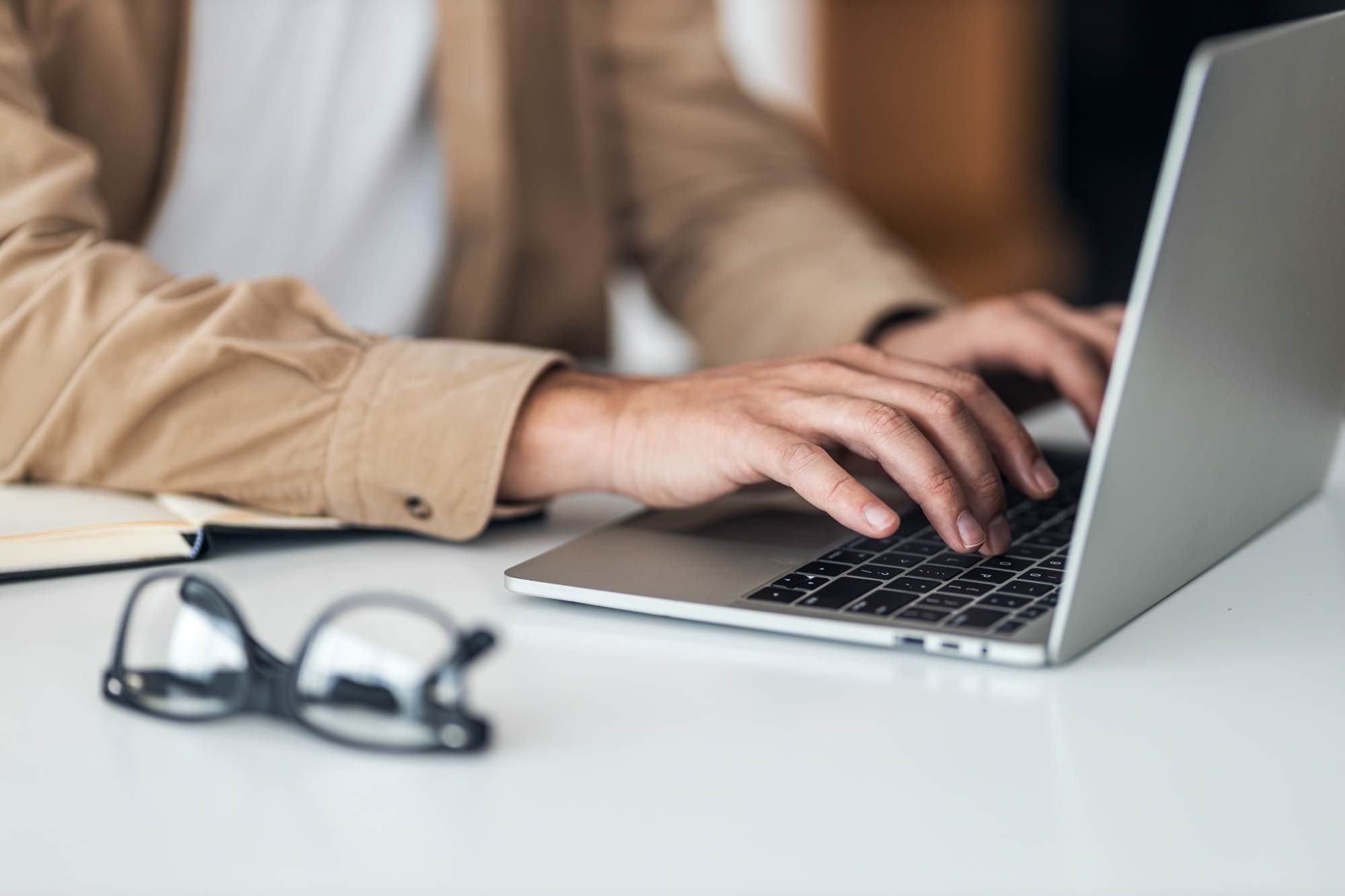 Person typing on a laptop at a desk with glasses and a notebook nearby.