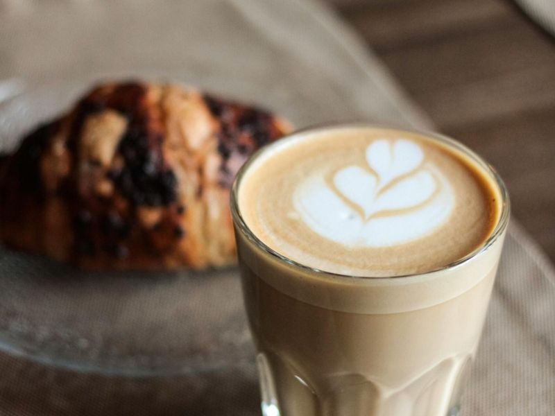 A glass of latte with heart latte art next to a chocolate croissant on a glass plate.