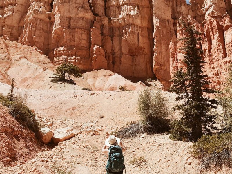 Person with a backpack hikes toward tall red rock formations under a blue sky with wispy clouds.
