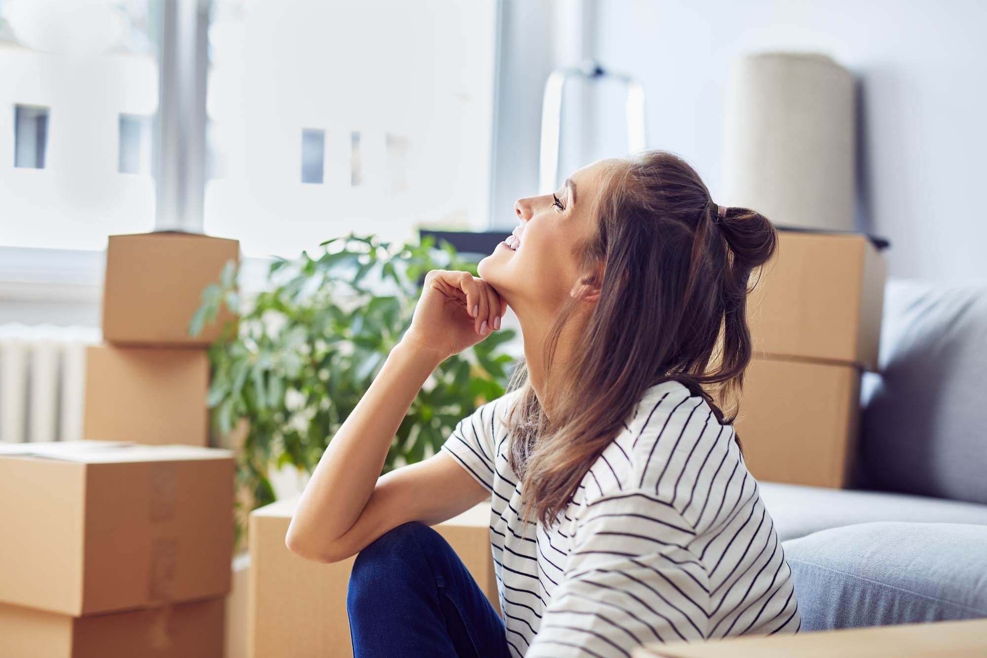 Woman sitting on the floor and smiling, surrounded by moving boxes in a bright living room.