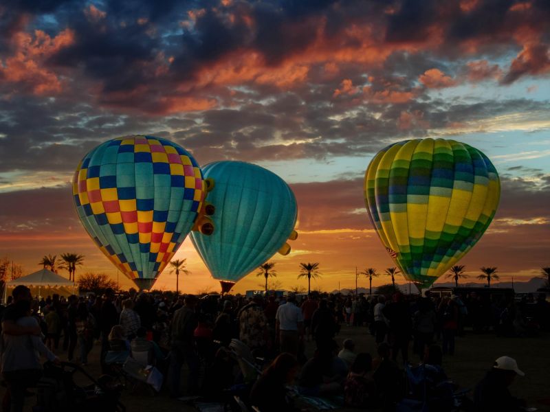 Three colorful hot air balloons above a crowd at sunset, with a dramatic sky and palm trees in the background.