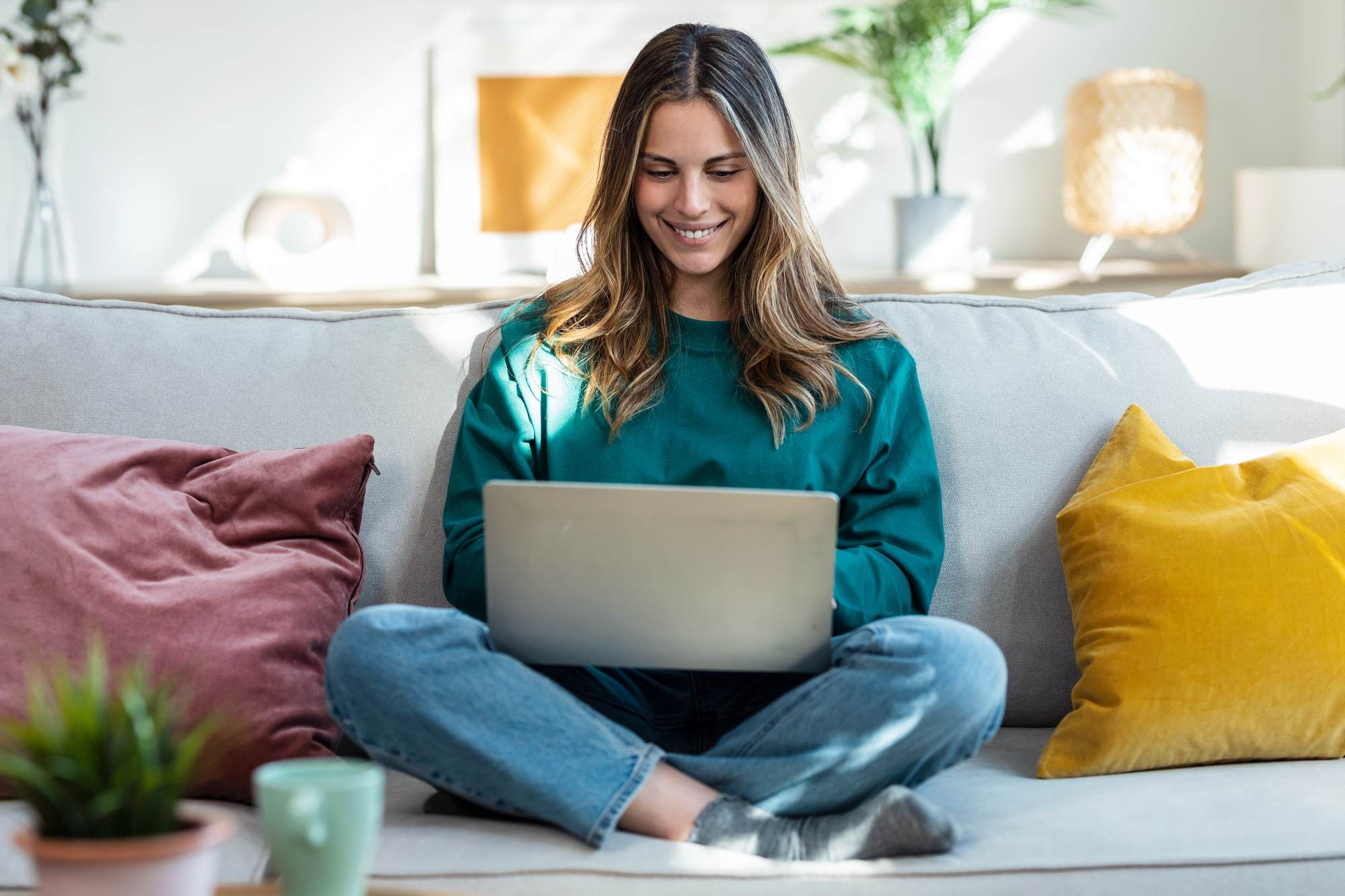 Woman sitting cross-legged on a sofa, smiling while using a laptop in a cozy, well-lit living room.