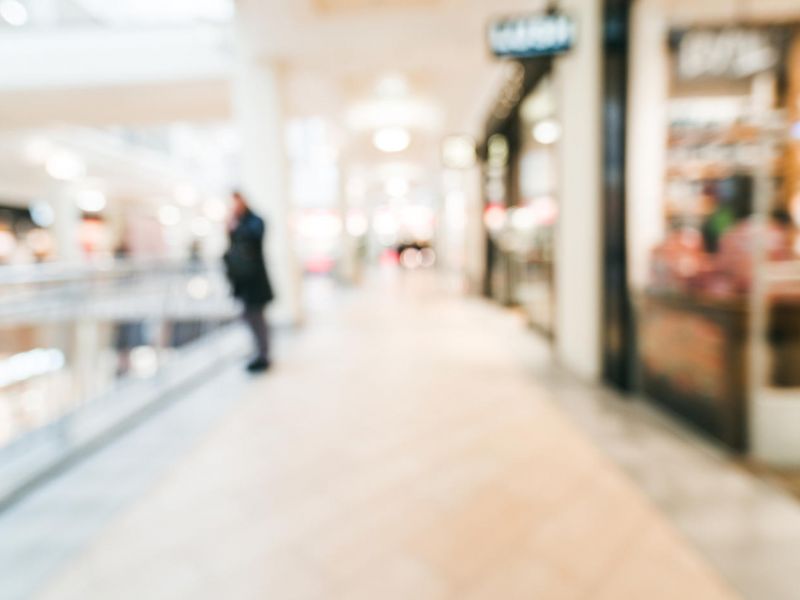 Blurry view of a person standing in a bright, modern shopping mall corridor with stores on each side.