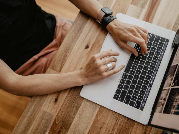 Person typing on a laptop at a wooden desk, with a smartwatch and rings visible on their hands.
